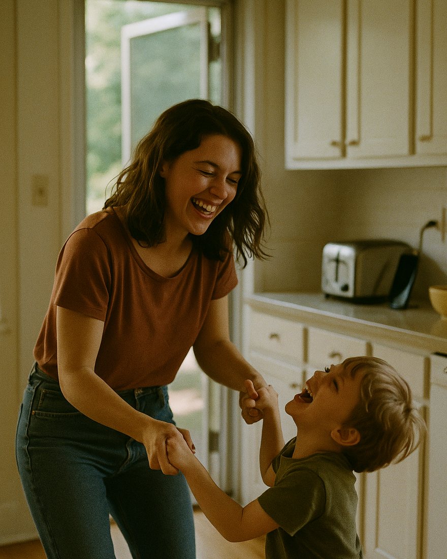 A mother and her small son laughing and dancing together in a bright American kitchen, white cabinets, the screen door open to a green yard behind them